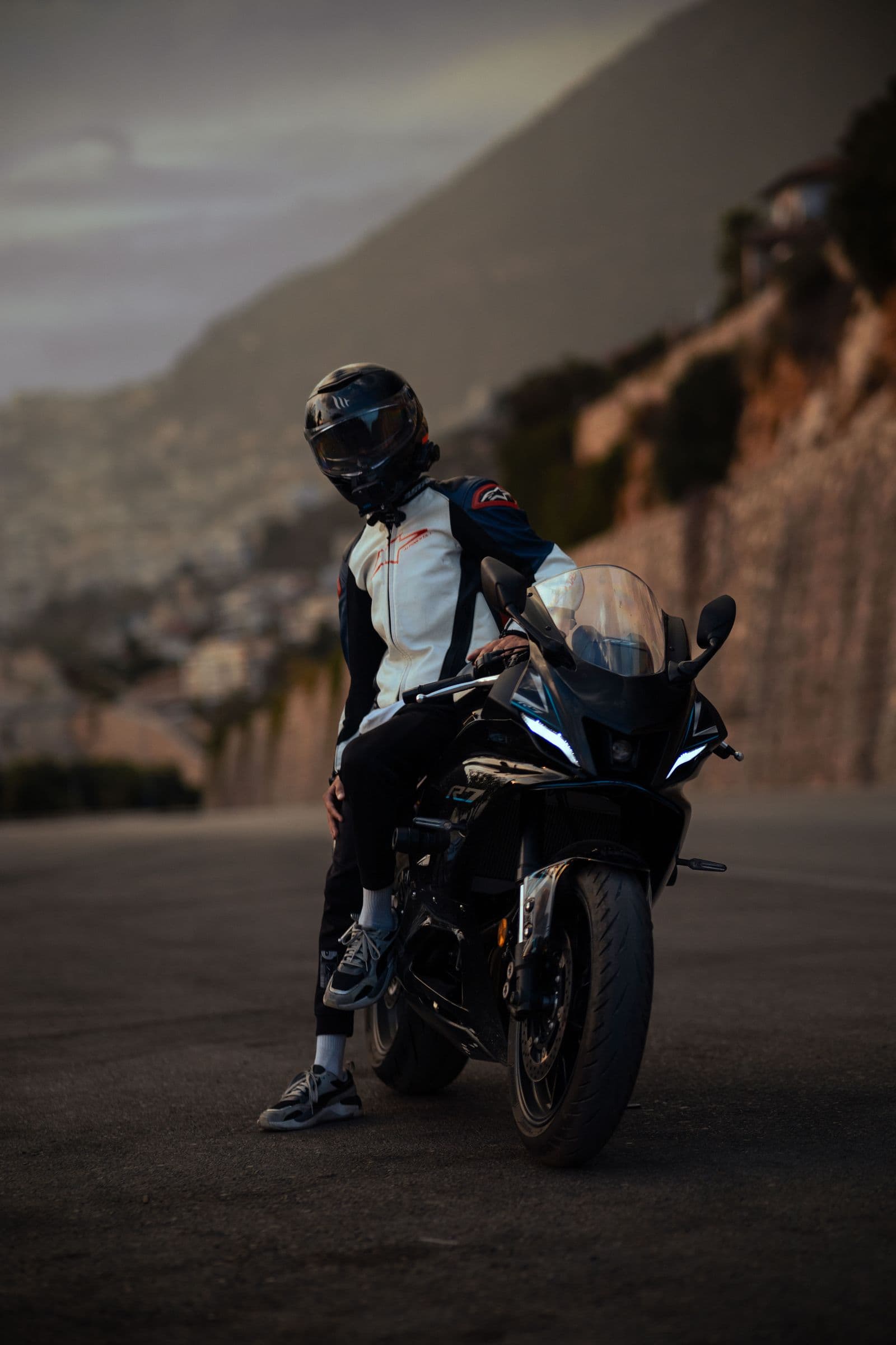 A rider in leathers leaning on a black sportsbike on a dusk mountain road
