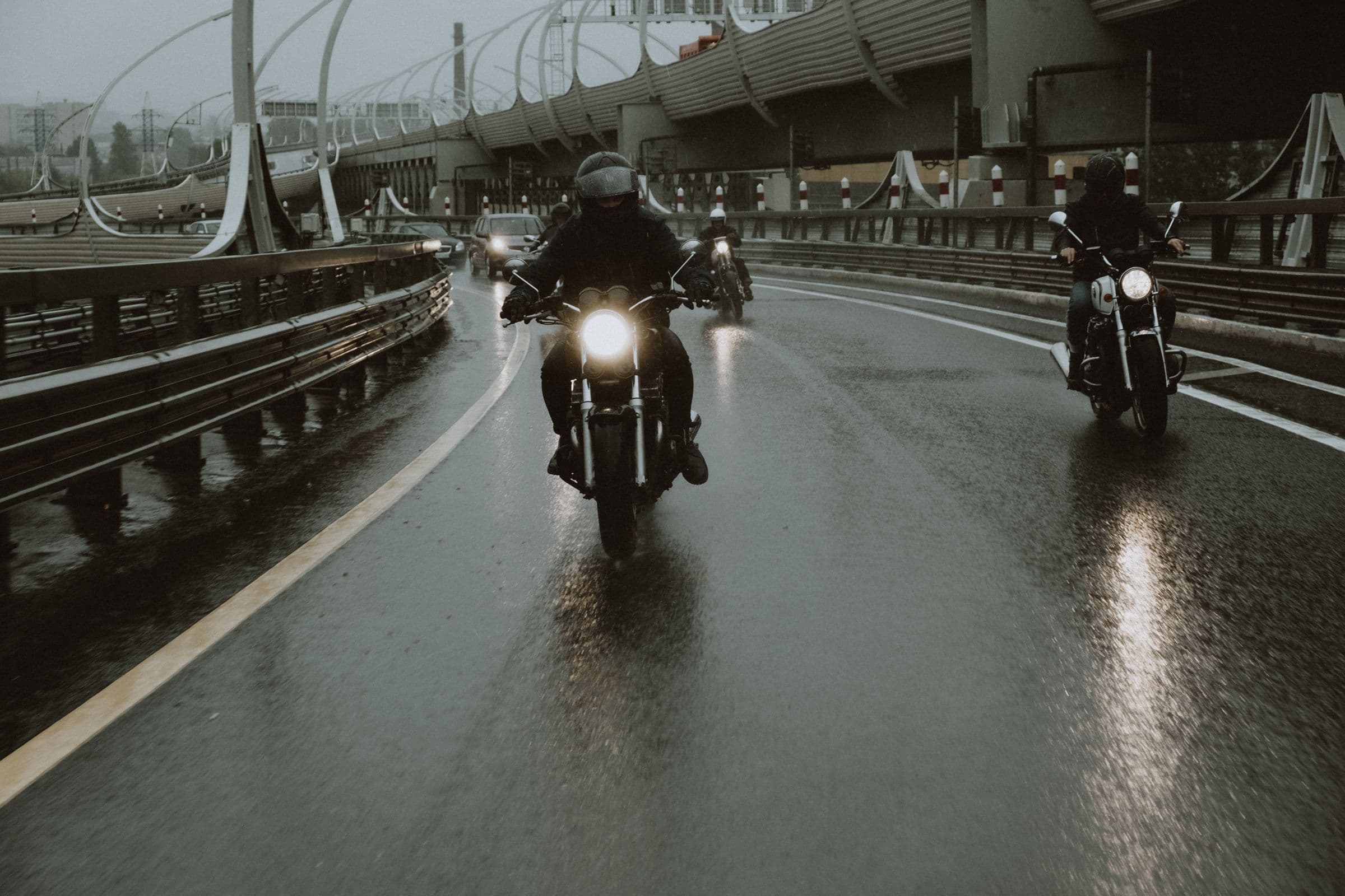 Three motorcyclists riding a rain-soaked highway under an overcast sky, headlights on